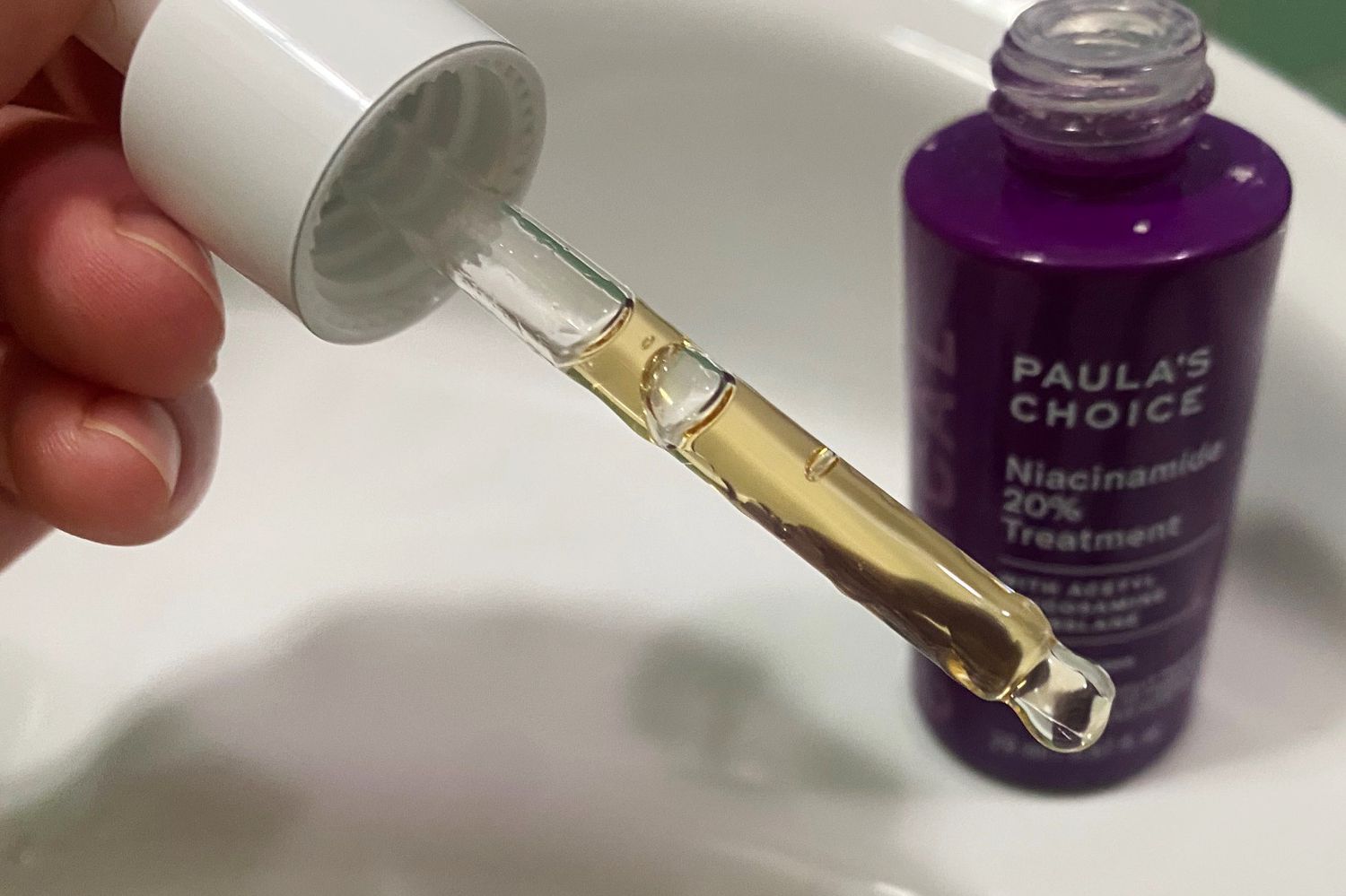 A hand holding the Paula's Choice Clinical Niacinamide 20% Treatment dropper near the bottle