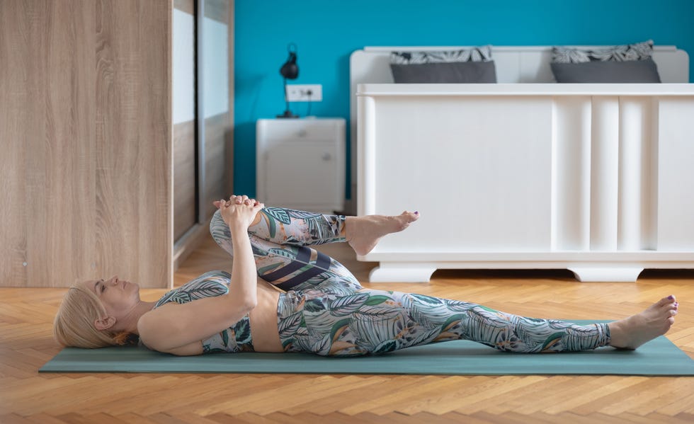 woman lying on mat at home and stretching leg bent in knee to chest