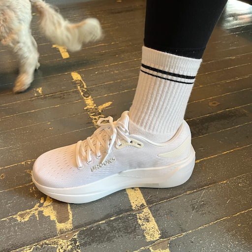 A person wearing a white running shoe and a striped sock, standing on a wooden floor.