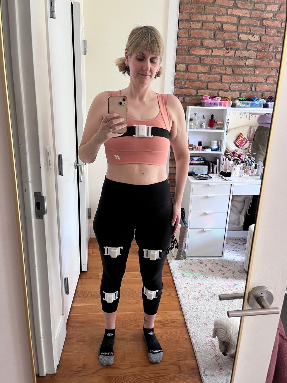 Person wearing a sports bra and fitted pants with attachments, standing in front of a mirror.