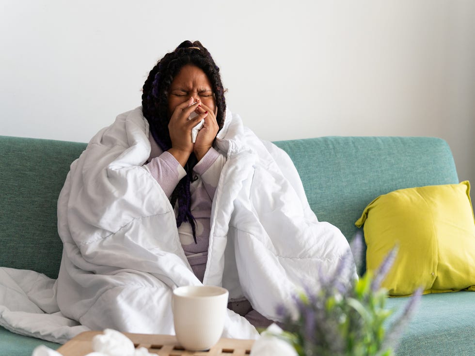 common pneumonia symptoms - young woman sitting on the sofa at home, covered with a blanket, blowing her nose, suffering from a cold or flu