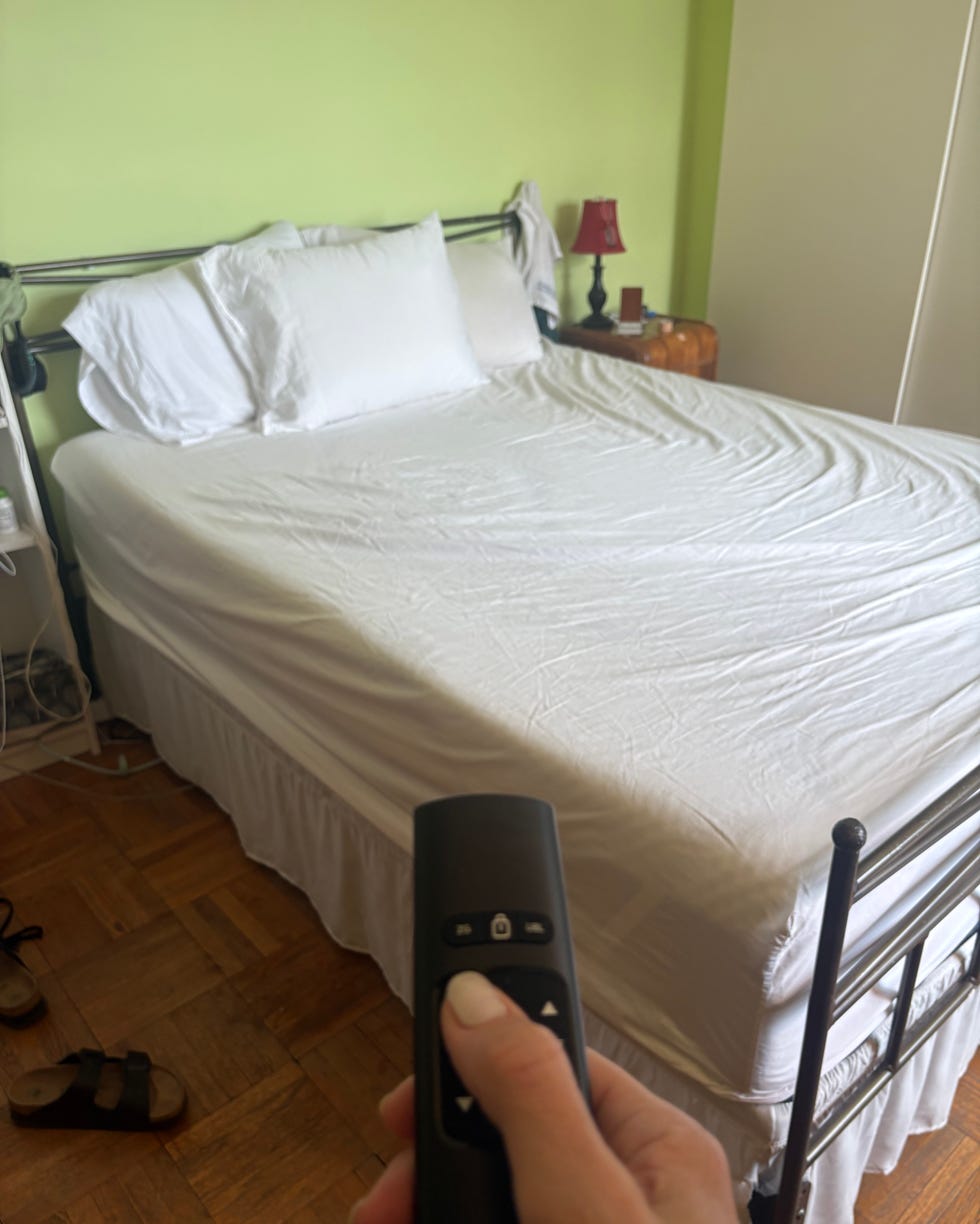 Hand holding remote control aimed at a neatly made bed with white linens in a bedroom.