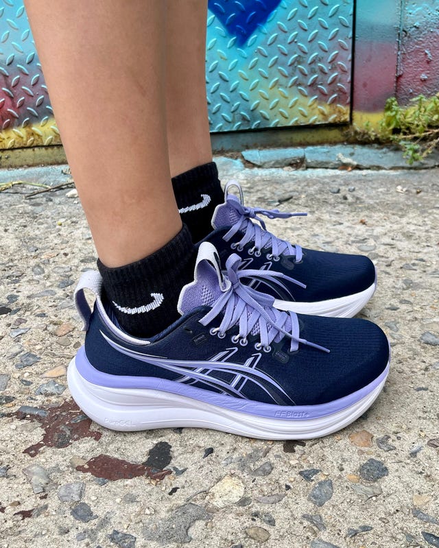 Purple and navy athletic shoes worn with black socks, standing on textured pavement with a colorful background.
