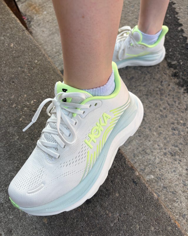 A close-up of a foot wearing white HOKA athletic shoes with neon green accents on a textured pavement surface. A close-up of a foot wearing white HOKA athletic shoes with neon green accents on a textured pavement surface.