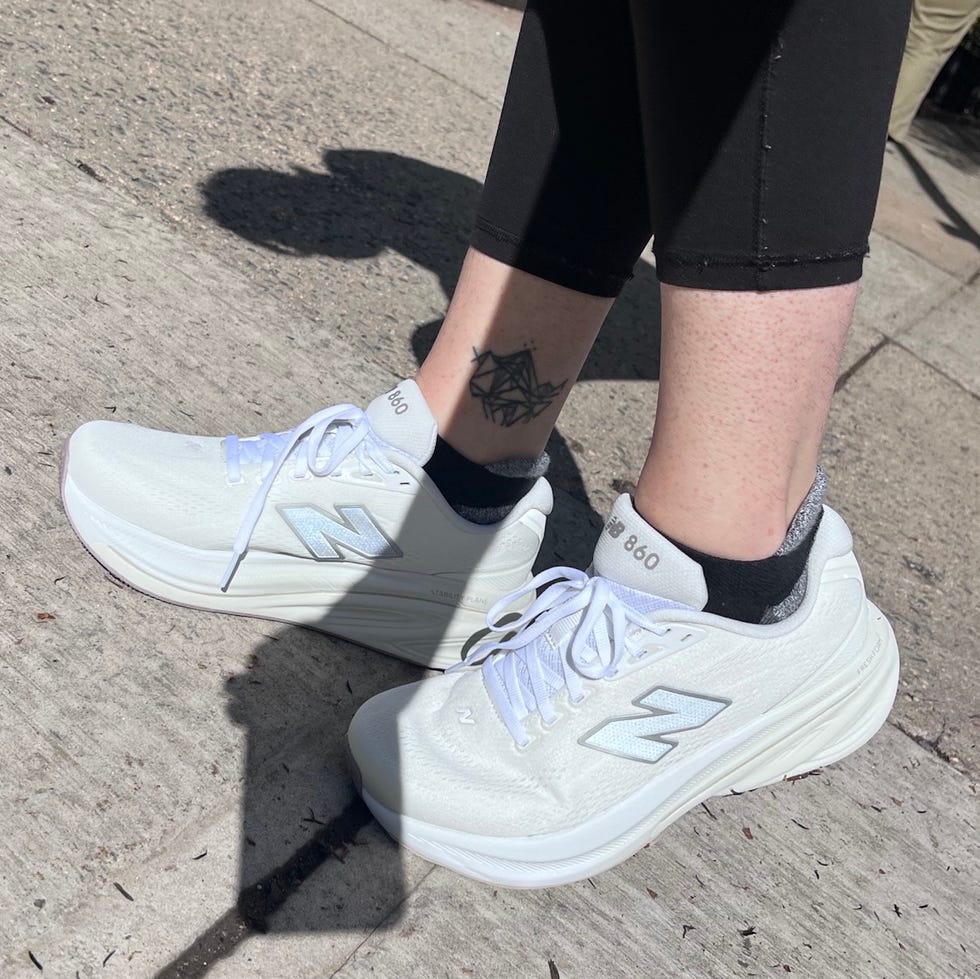 White New Balance sneakers worn on a person’s feet, with a visible tattoo on the ankle, standing on a sidewalk.