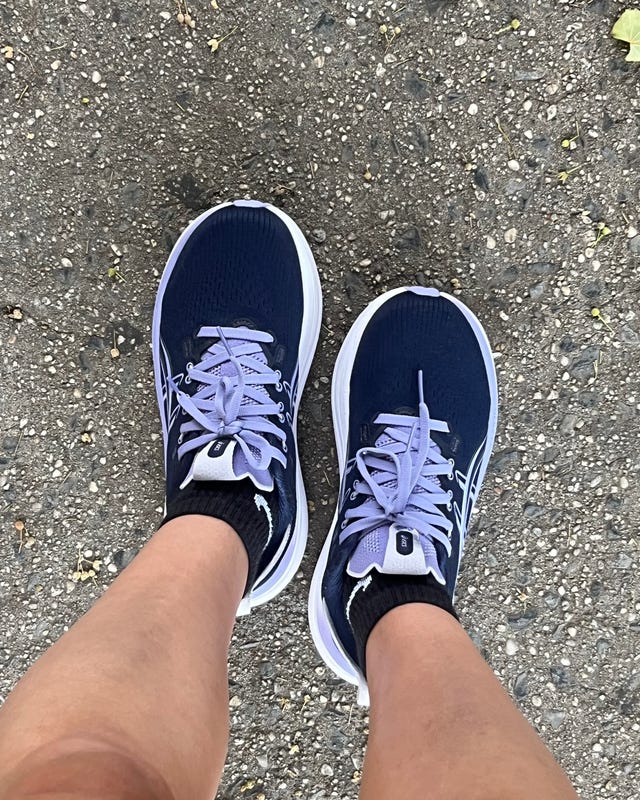 A pair of navy running shoes on a person's feet, standing on a gravel path with scattered leaves.