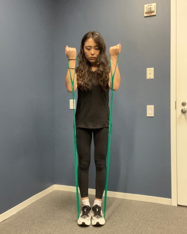 Person demonstrating an exercise using a green resistance band, standing on a gray floor with a blue wall in the background.