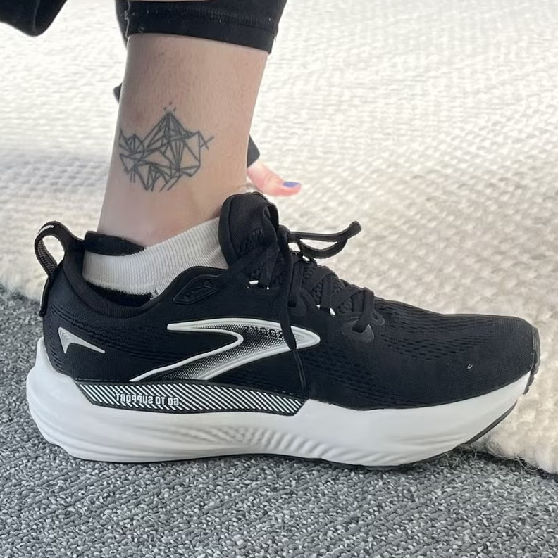 A person adjusting a black athletic shoe while showing off a geometric tattoo on their ankle.