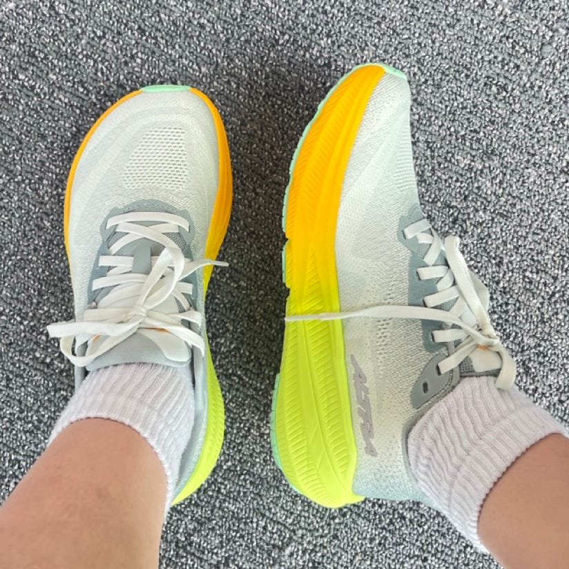 Athletic shoes featuring a light gray upper and bright yellow soles, paired with white socks on a textured floor.