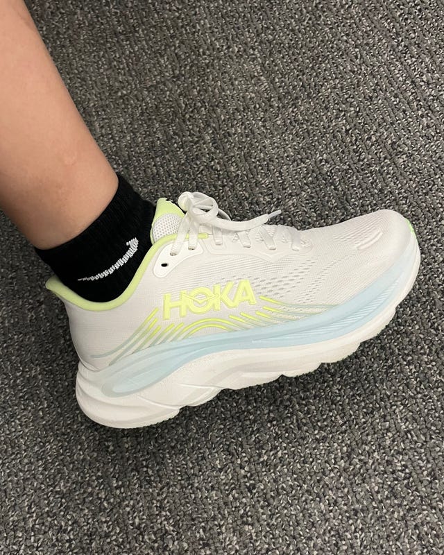 White athletic shoe with neon accents worn with a black sock, resting on a gray carpeted surface. White athletic shoe with neon accents worn with a black sock, resting on a gray carpeted surface.