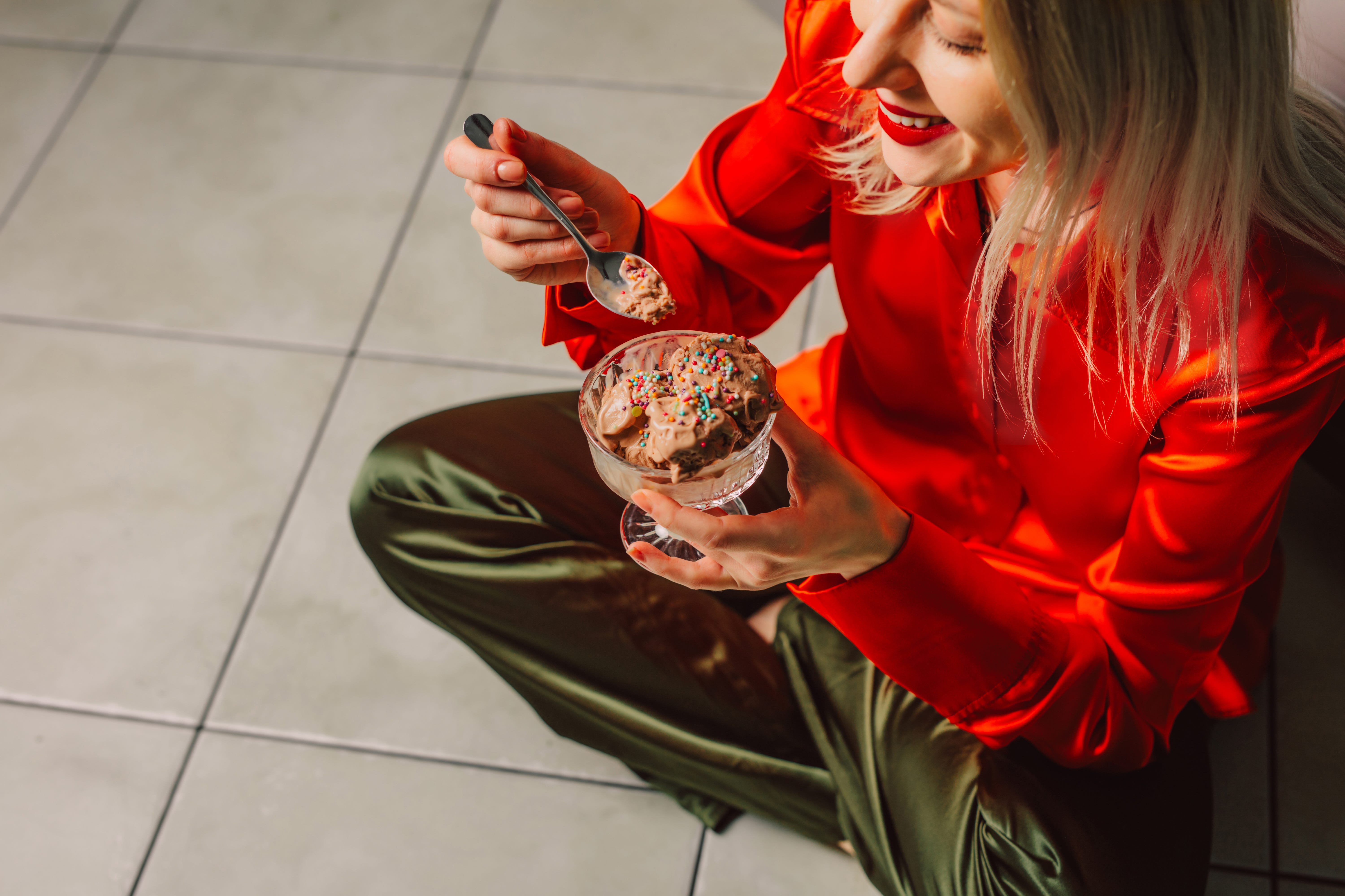 Woman with blonde hair eating ice cream on kitchen floor in stylish attire