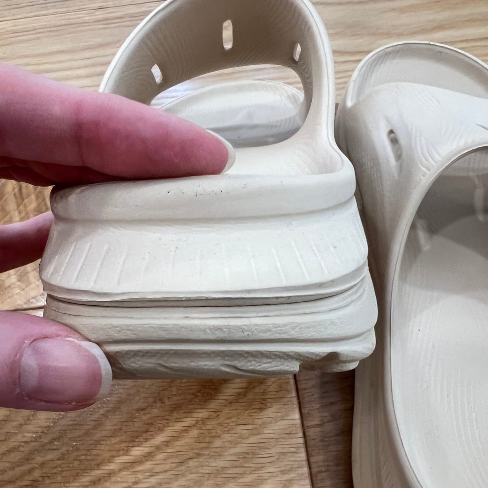 Hand holding a light-colored shoe with a thick sole, resting beside another similar shoe on a wooden surface.