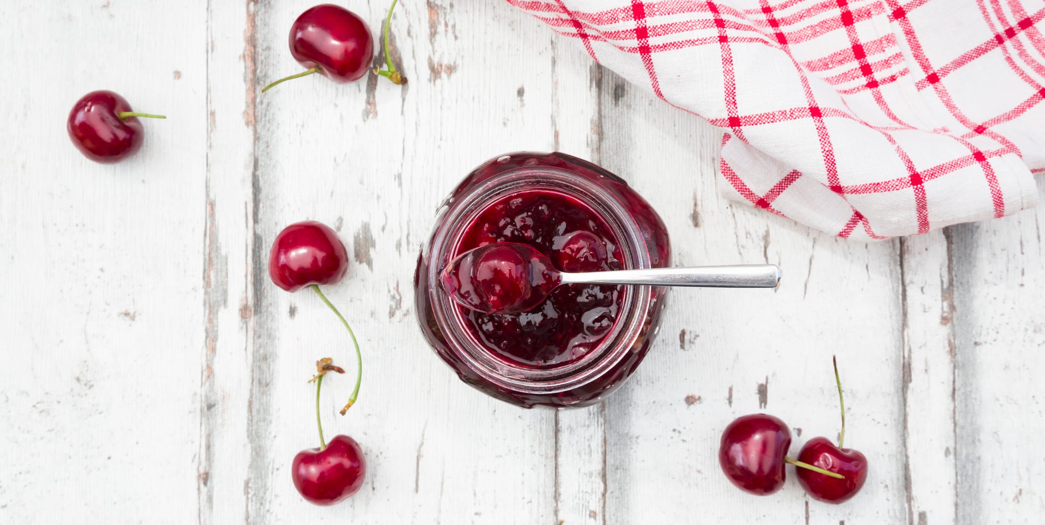Cherries beside open jar of cherry jam on white wood