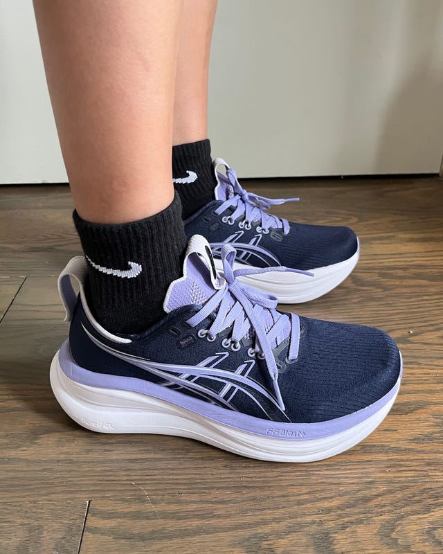 Close-up of a person wearing navy blue athletic shoes with purple accents and black socks on a wooden floor.
