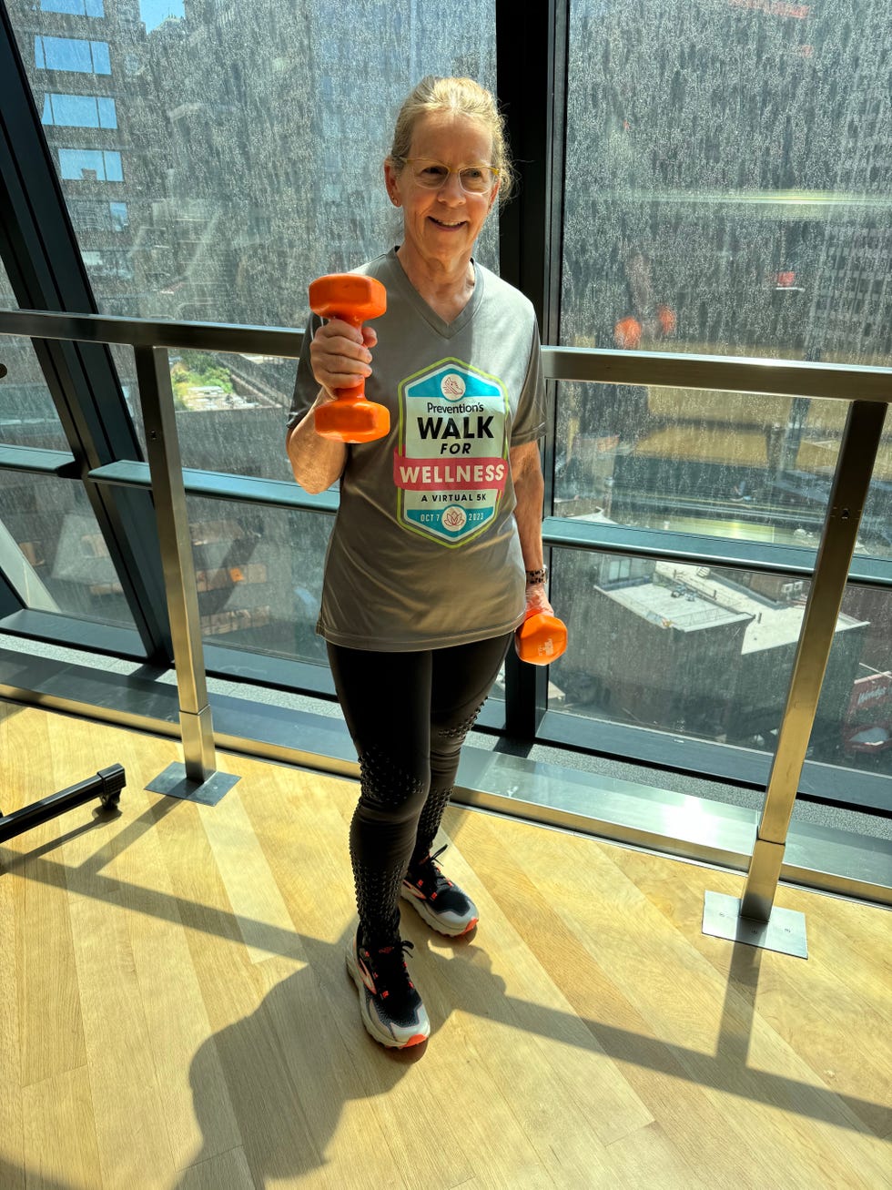 Older woman in fitness attire holding a small orange dumbbell indoors with a city view in the background.
