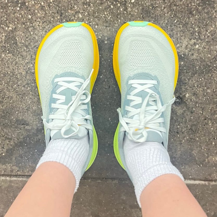 Aerial view of light blue athletic shoes with yellow and green accents, worn with white socks on a concrete surface.