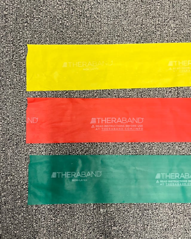 Three Theraband resistance bands in yellow, red, and green laid out on a textured gray surface.