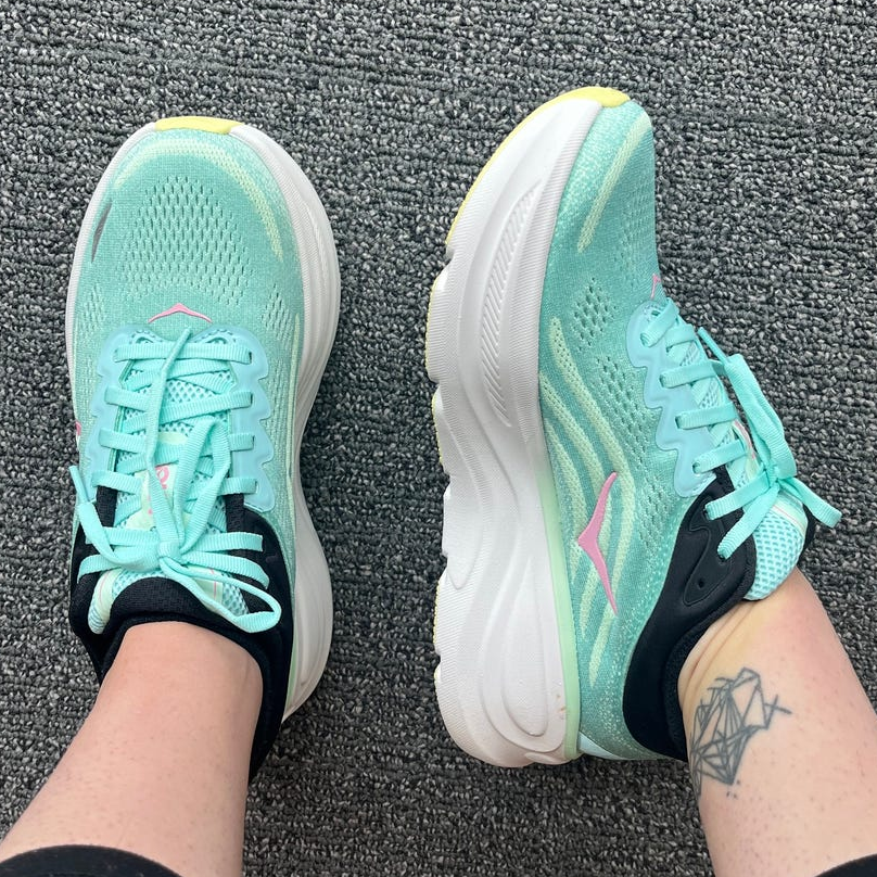 Pair of turquoise athletic shoes with black accents, worn by a person sitting on a textured surface.