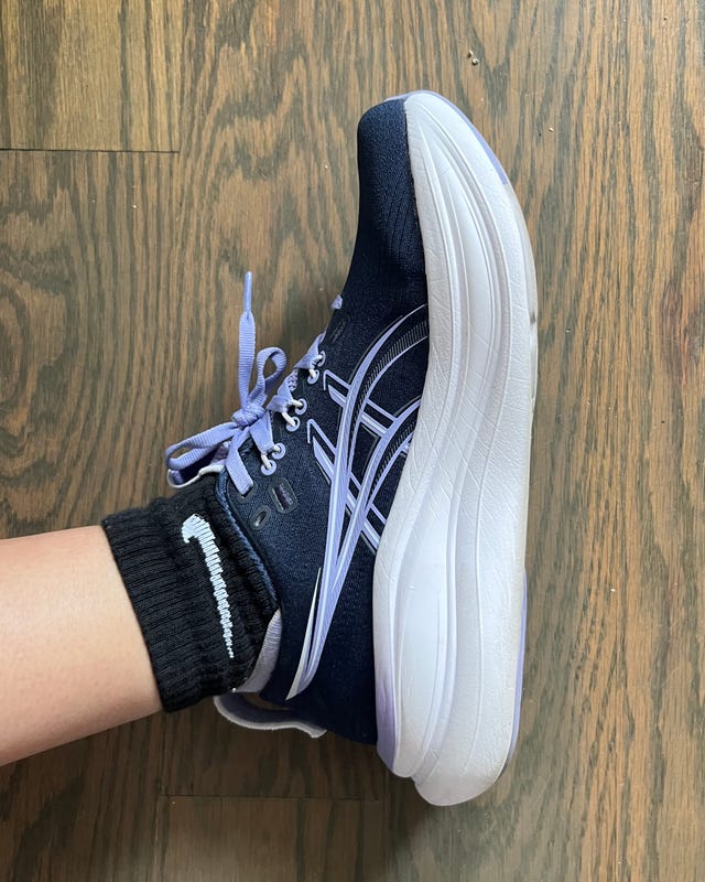 Blue athletic shoe on a foot wearing a black Nike sock, positioned on a wooden floor.