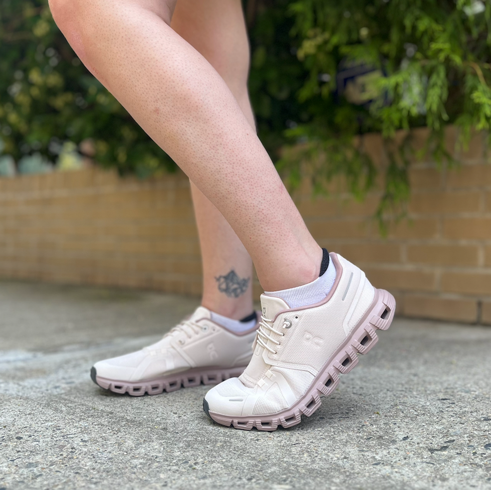 Light pink athletic shoes on a person's foot with a tattoo visible on the leg, standing on a concrete surface.