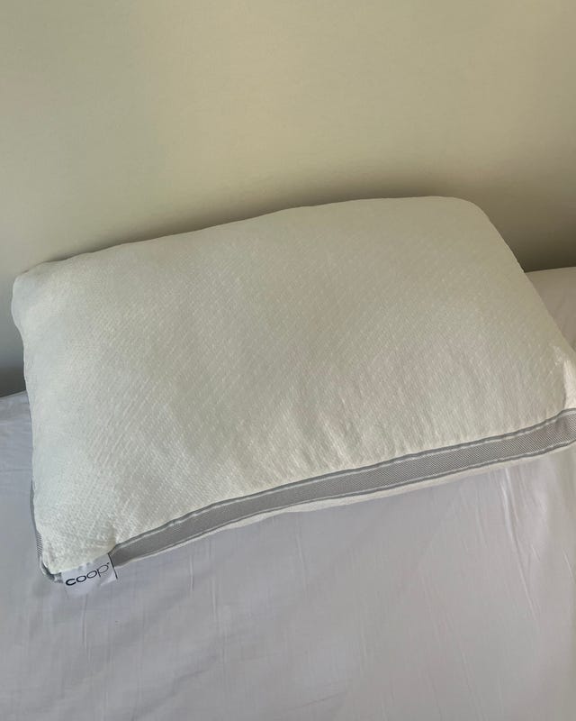 A soft, white pillow resting on a light-colored bed sheet against a neutral wall.