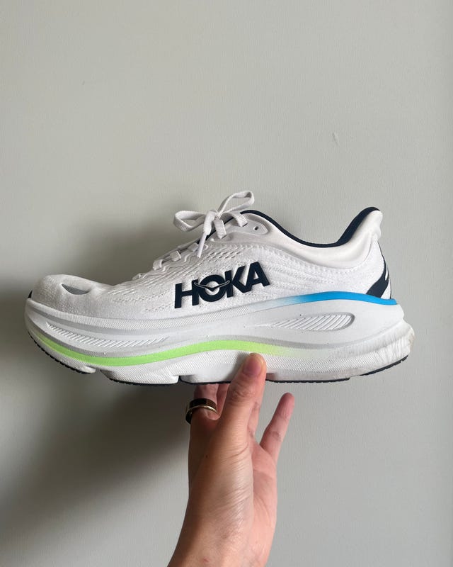 White HOKA running shoe held against a light-colored wall.