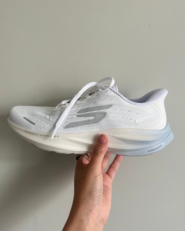 A white athletic shoe held against a neutral wall background. A white athletic shoe held against a neutral wall background.