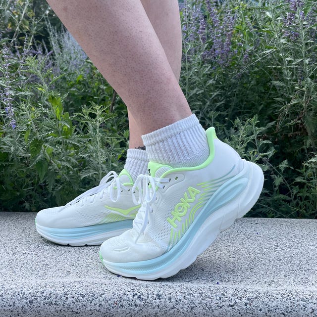 White HOKA running shoes with neon accents, positioned on a concrete step against a backdrop of greenery.