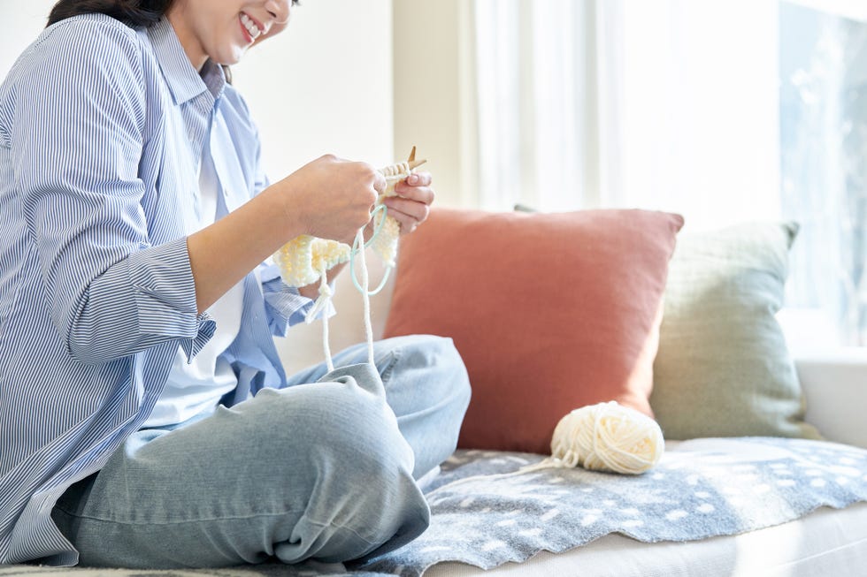 A person knitting comfortably on a couch, surrounded by colorful pillows and a ball of yarn.