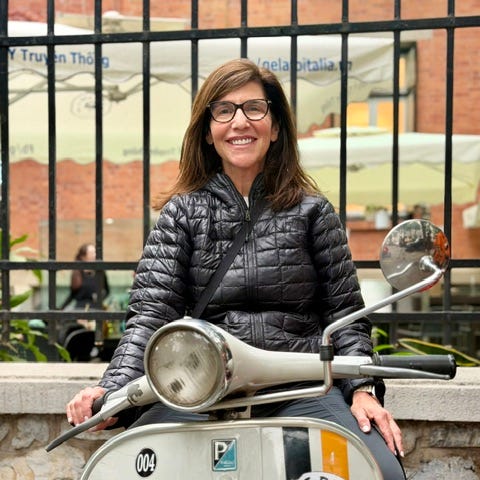 A woman with glasses smiles while sitting on a vintage Vespa scooter in a lively outdoor setting.