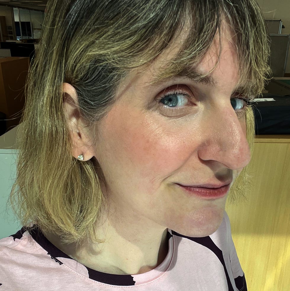 A person with shoulder-length hair wearing a pink shirt with a black design wearing laura gellar wonder balm blush