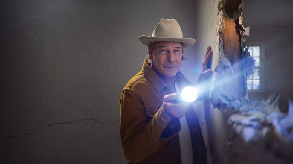 A person in a cowboy hat holds a flashlight, examining a wall with visible damage in a dimly lit room.