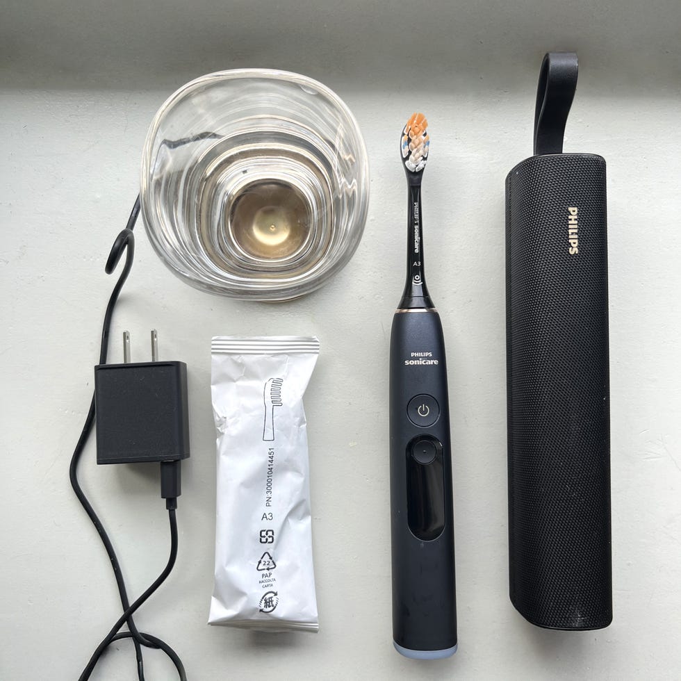 A Philips Sonicare toothbrush, charger, replacement brush head, glass, and travel case on a light surface.