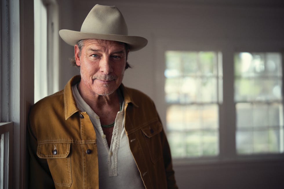 Man in a cowboy hat and a mustard-colored jacket leaning against a wall, smiling softly in a well-lit room.