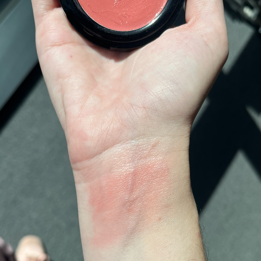 Swatch of blush on a hand with a hint of color on the wrist, with a background showing a shoe and pavement.
